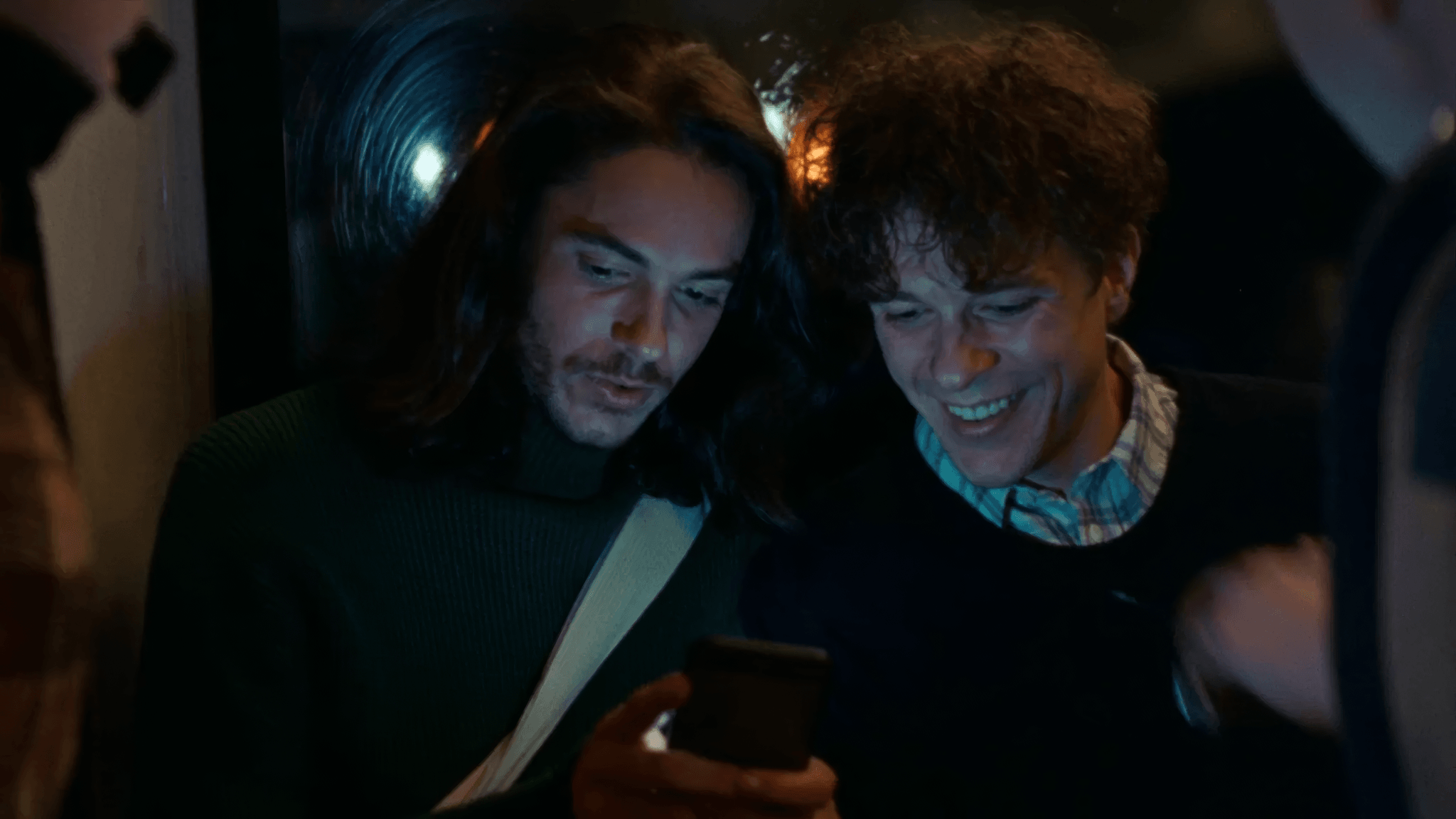 Two friends leaning in over one phone at night, faces lit by the screen, one grinning