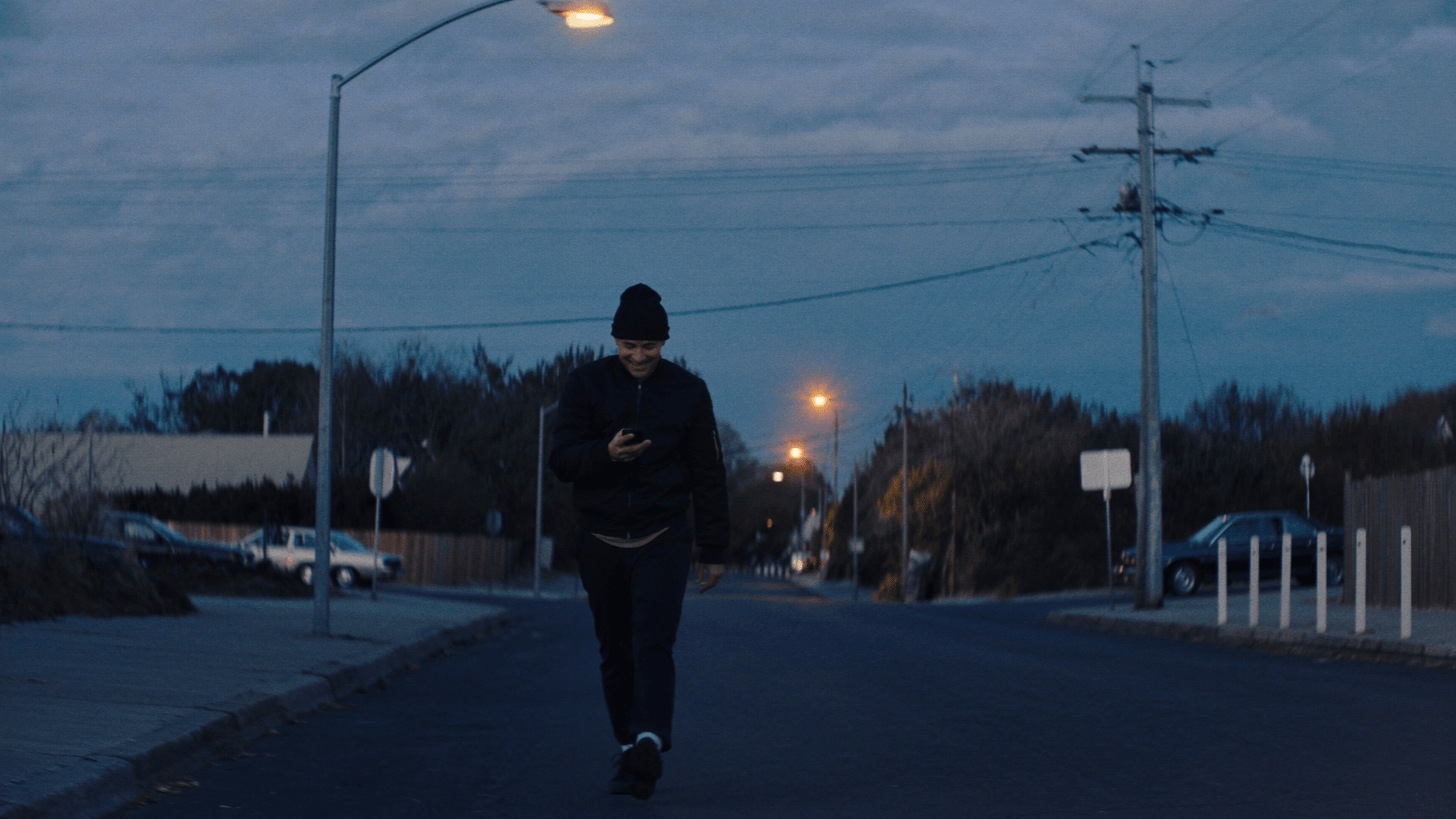 A young man in a beanie walking down an empty street at dusk, smiling at his phone under the streetlights